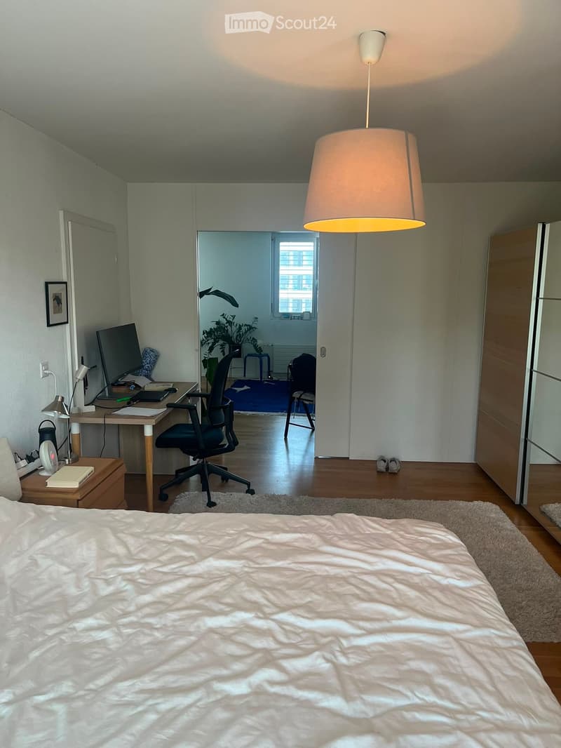 Modern bedroom with a large bed, desk with a computer, office chair, hanging lamp, and a view into an adjacent room with a plant and stool.