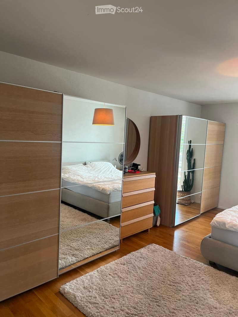 Modern bedroom with wooden flooring, large mirrored wardrobe, bed with white bedding, nightstand with lamp, and a round mirror above it.