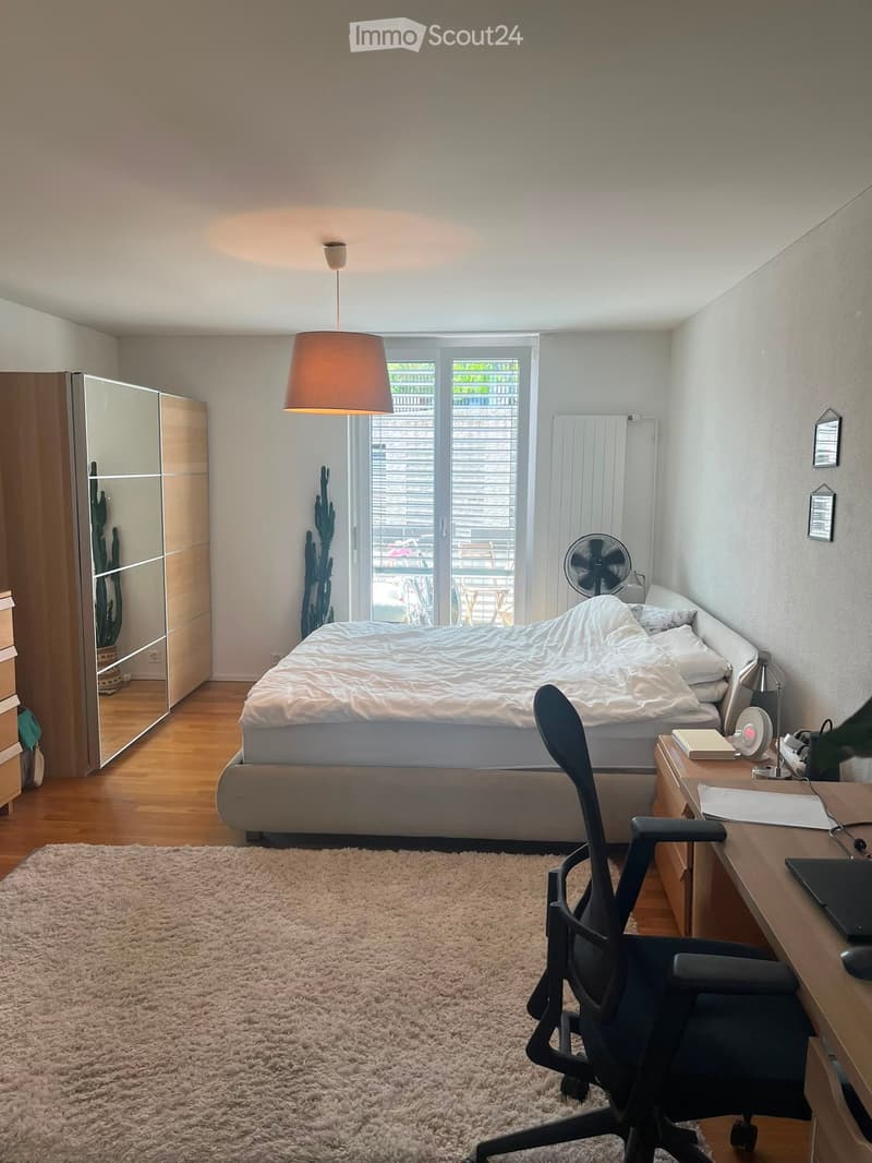 Modern bedroom with a large bed, desk, chair, and a fan. Features a large window with blinds, a hanging lamp, and a carpeted floor.