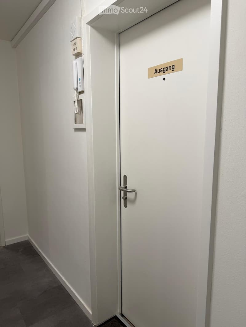 A narrow hallway with white walls, a closed door labeled "Ausgang" (exit), and a wall-mounted intercom system.