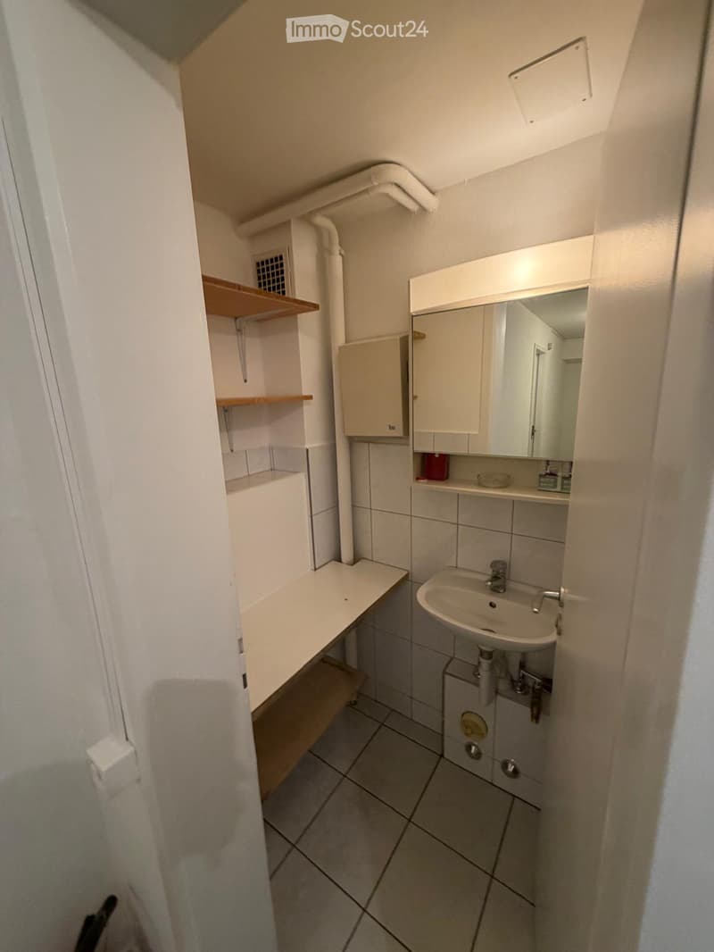 Small bathroom with tiled floor, white walls, a sink with a mirror cabinet above, and a wooden shelf unit on the left side.