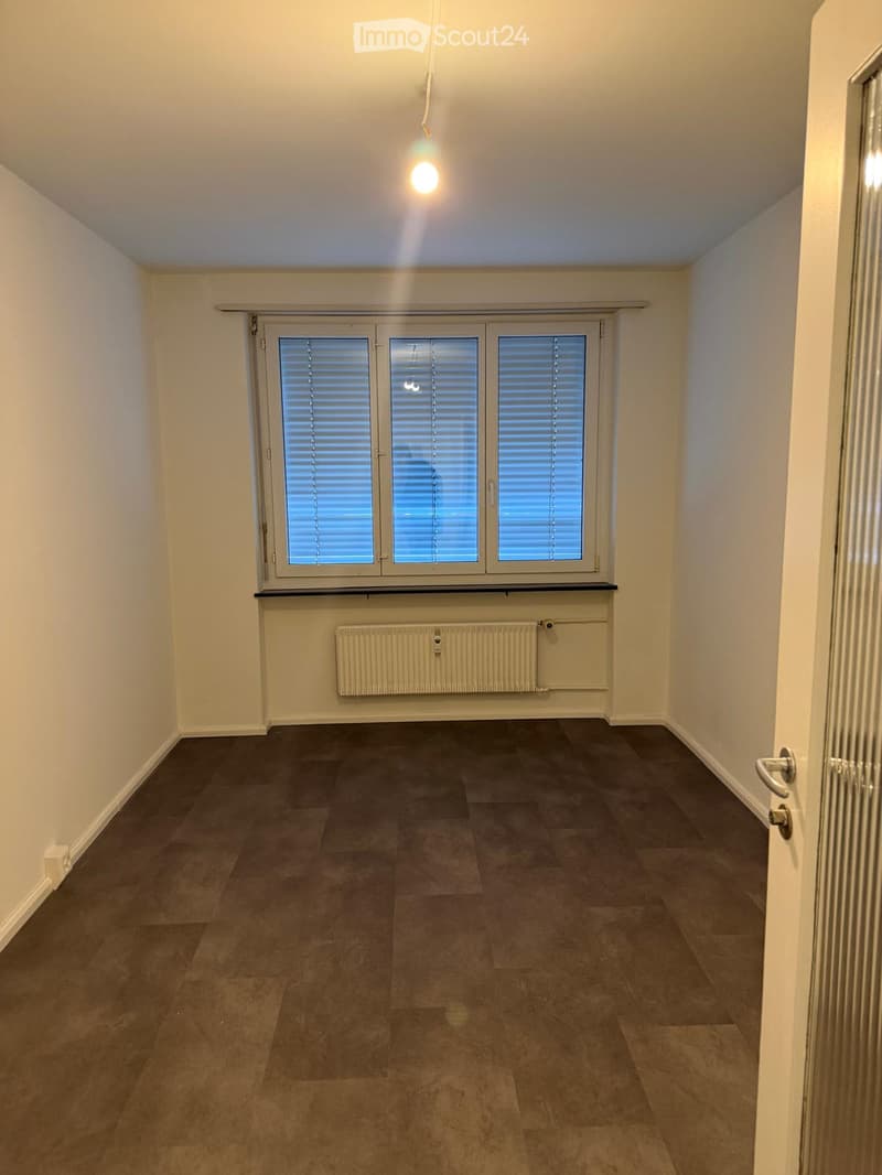 Empty room with beige walls, dark tiled floor, large window with blinds, radiator, and a door.