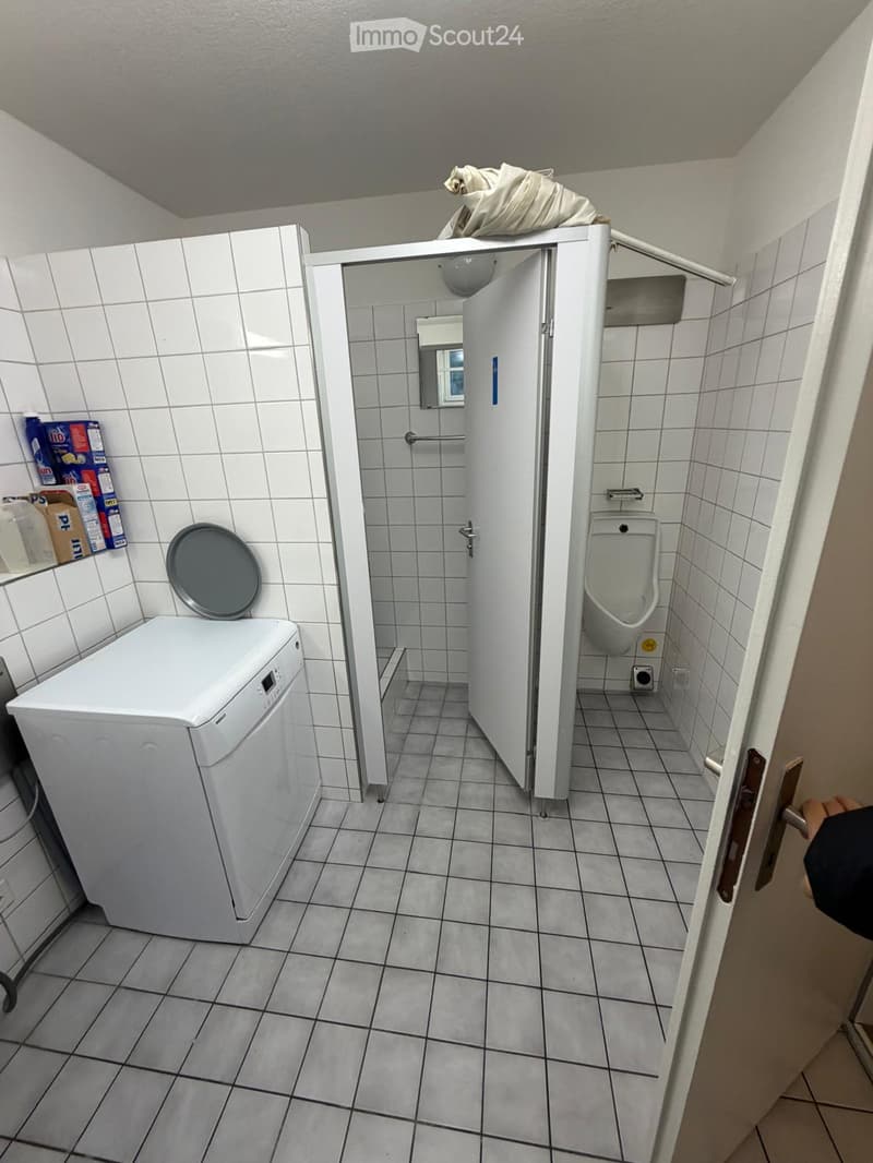 Bathroom with a washing machine, urinal, mirror, and a small storage area with boxes and cleaning supplies.