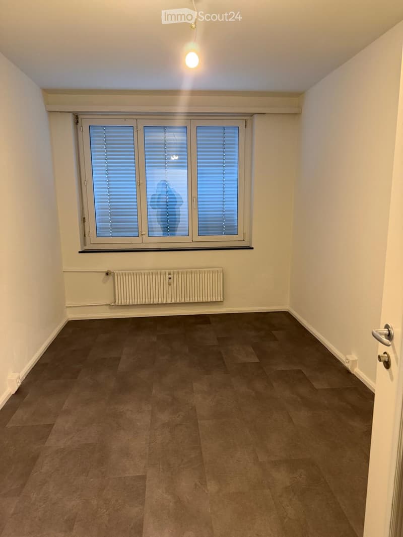 Empty room with wooden flooring, white walls, large window with blinds, radiator, and a single door.