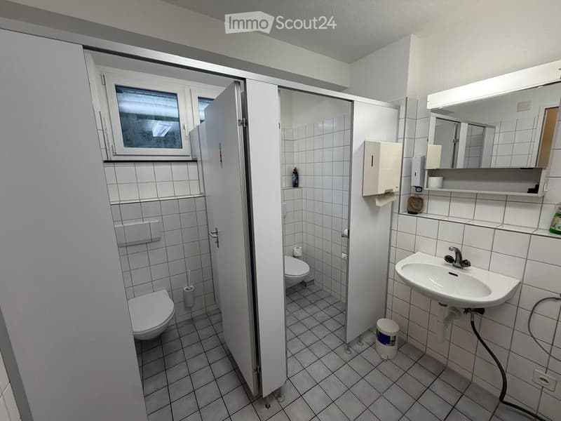 Modern bathroom with white tiles, a sink, toilet, mirror, and a separate shower stall. Includes a hand soap dispenser and a small trash bin.