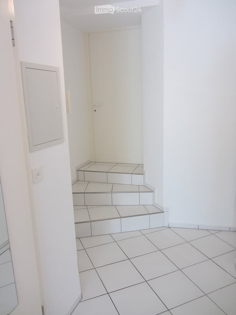 Empty hallway with tiled floor and white walls, featuring a small step-up section and electrical outlet.