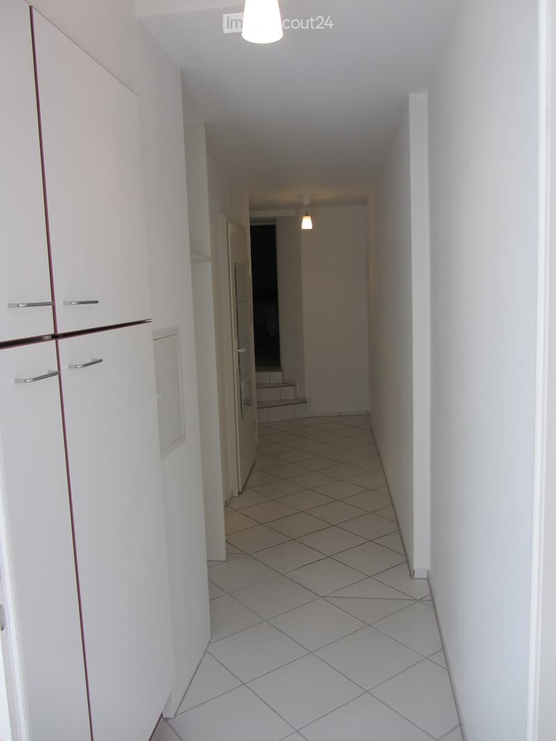 A clean, well-lit hallway with white tiled flooring and white walls. The hallway features a built-in white cabinet with silver handles on the left side and a staircase leading upwards in the background.