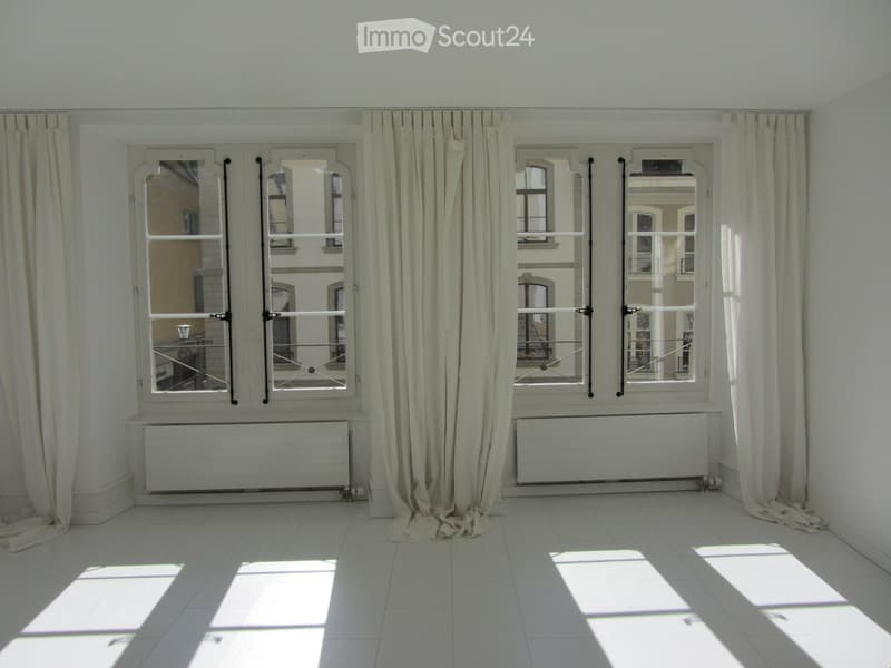 Empty room with white tiled floor, large windows with beige curtains, radiators below the windows, and natural light streaming in.