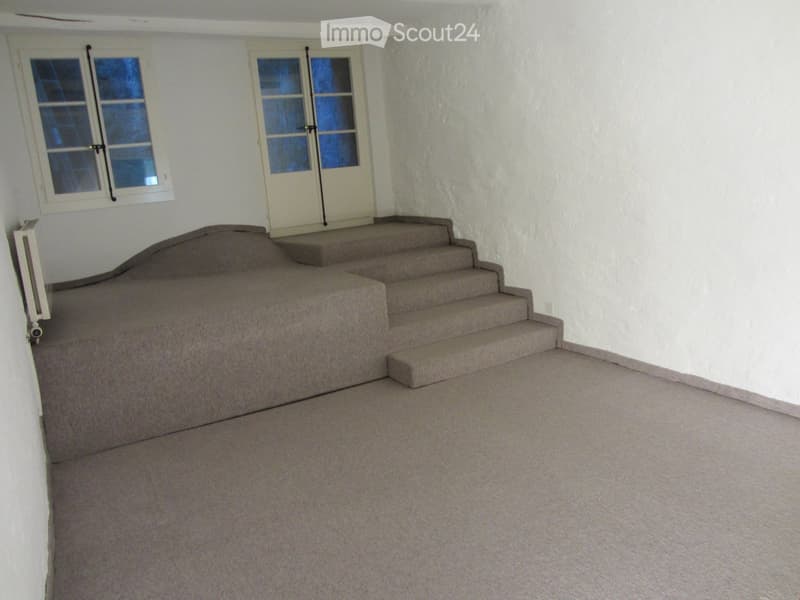 Empty room with carpeted floor, white walls, and a staircase leading upwards. Two windows and a door are visible on the back wall.