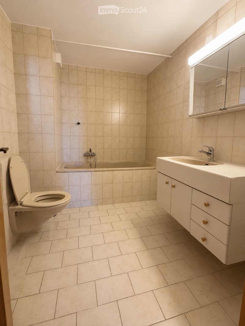 Modern bathroom with beige tiled walls and floor, featuring a toilet, bathtub, sink with mirror, and white cabinet.