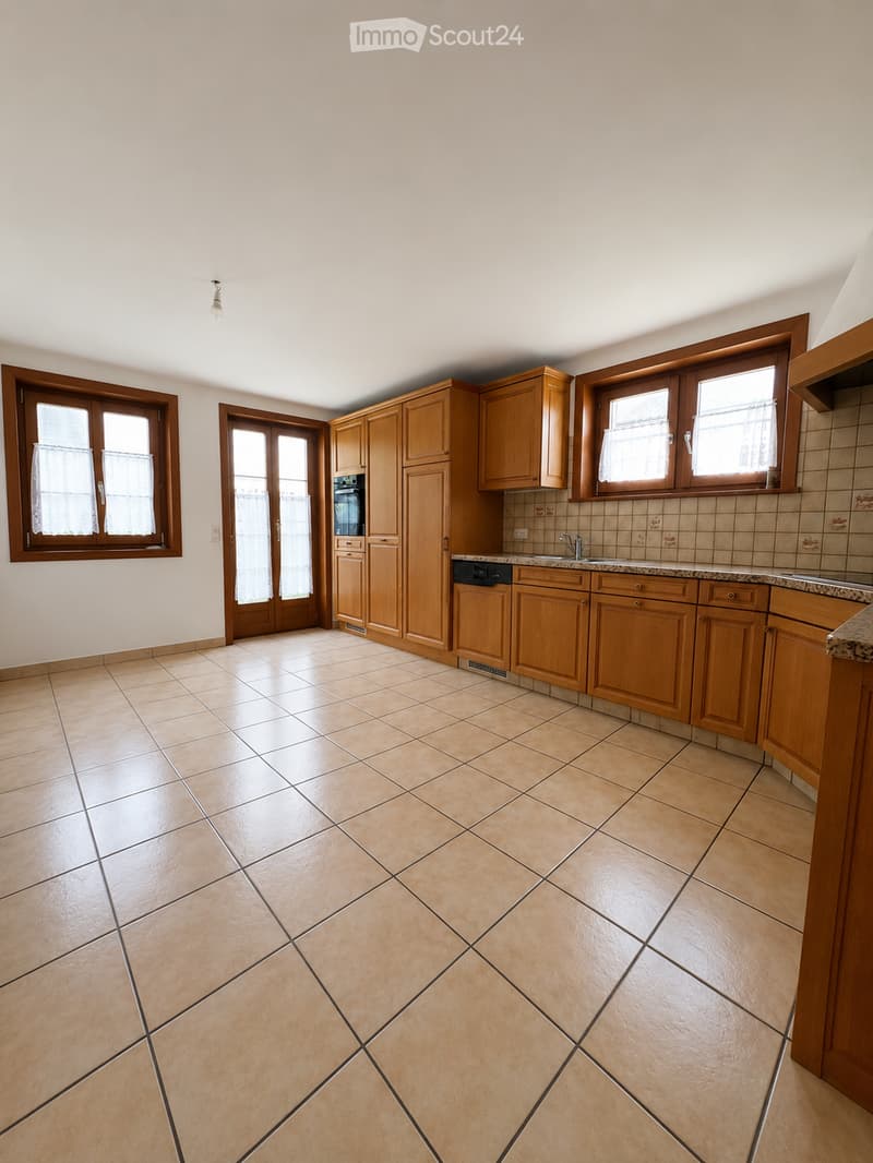 Empty kitchen with wooden cabinets, tiled floor, and large windows. No appliances or furniture are present.
