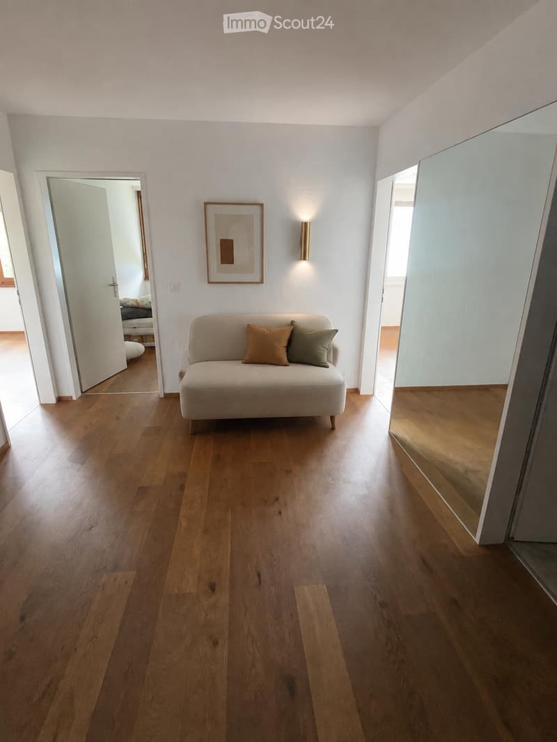 Living room with light wooden flooring, a beige sofa with two throw pillows, a wall-mounted picture frame, and a wall-mounted light fixture. The room features a large mirror on one wall and an open doorway leading to another room.