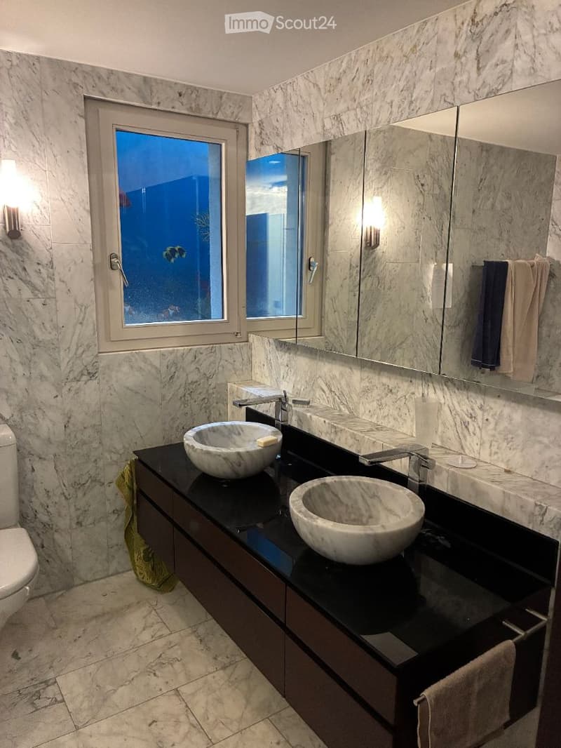 Modern bathroom with marble walls and floors, double sink vanity with integrated sinks, large mirror, and wall-mounted lighting.