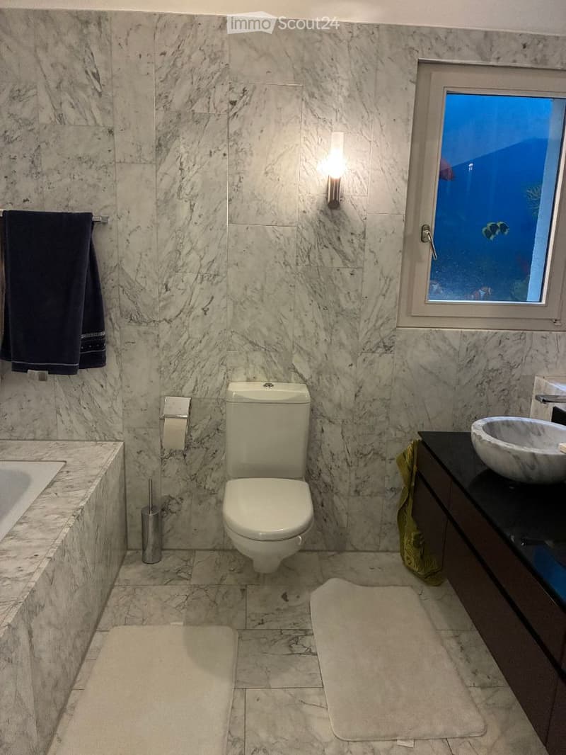 Modern bathroom with marble walls and floors, featuring a toilet, sink, bathtub, and a window with a scenic view.