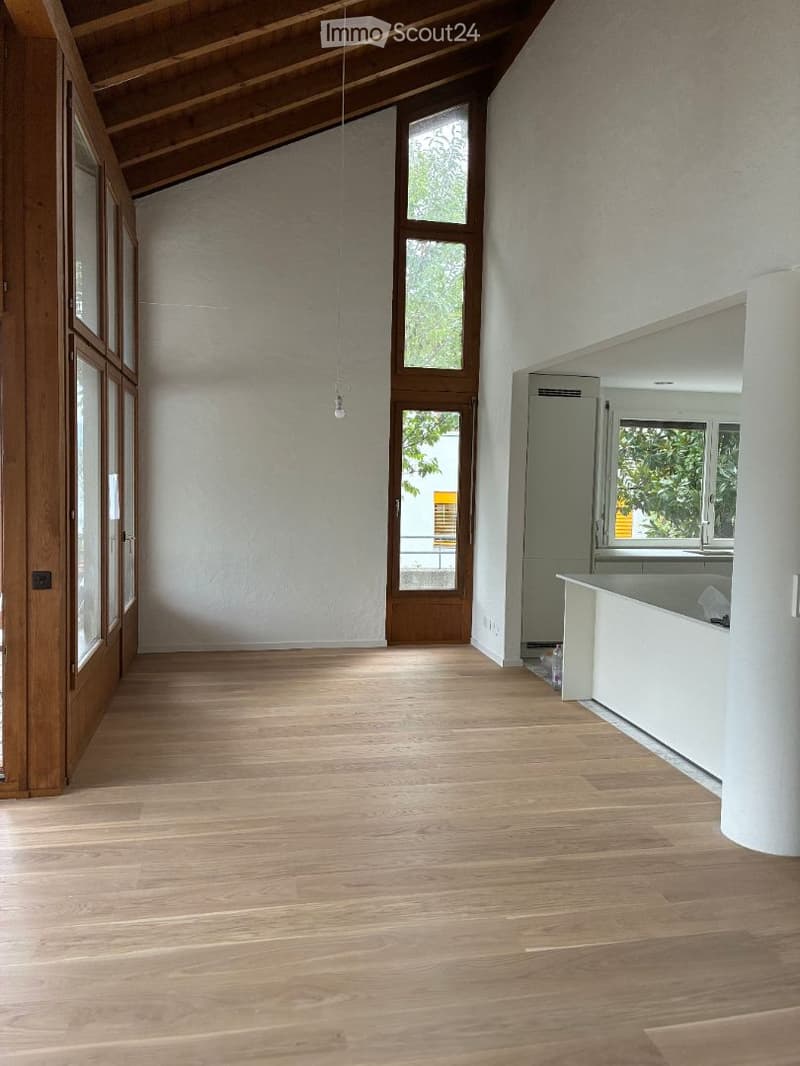 Bright, empty room with wooden flooring, white walls, large windows, and a visible kitchen area in the background.