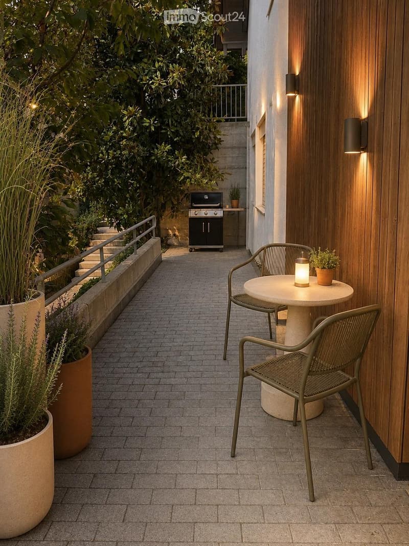 A modern outdoor balcony with a round table, two chairs, a small lamp, and a barbecue in the background, surrounded by greenery and a paved pathway.