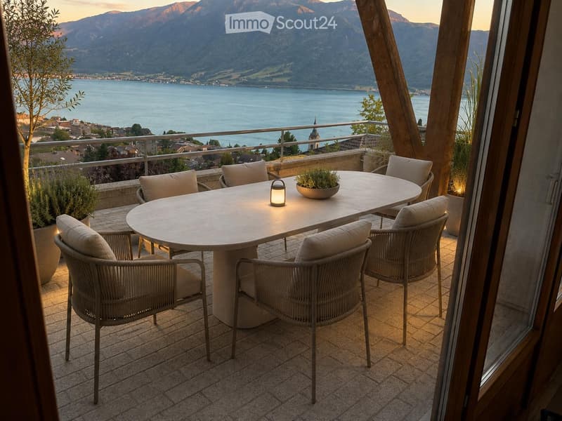 Balcony with a round table, six chairs, a small lamp, and potted plants, overlooking a lake and mountains.