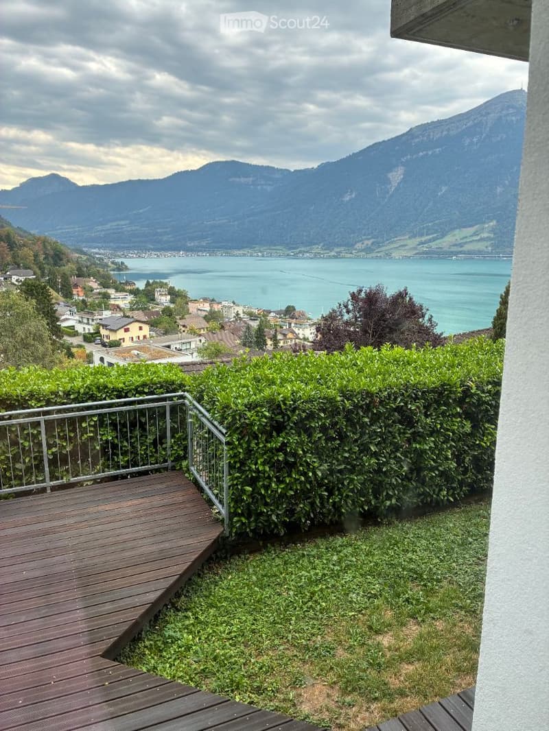 Balcony with wooden decking, metal railing, and lush green hedges overlooking a scenic lake and mountain landscape.