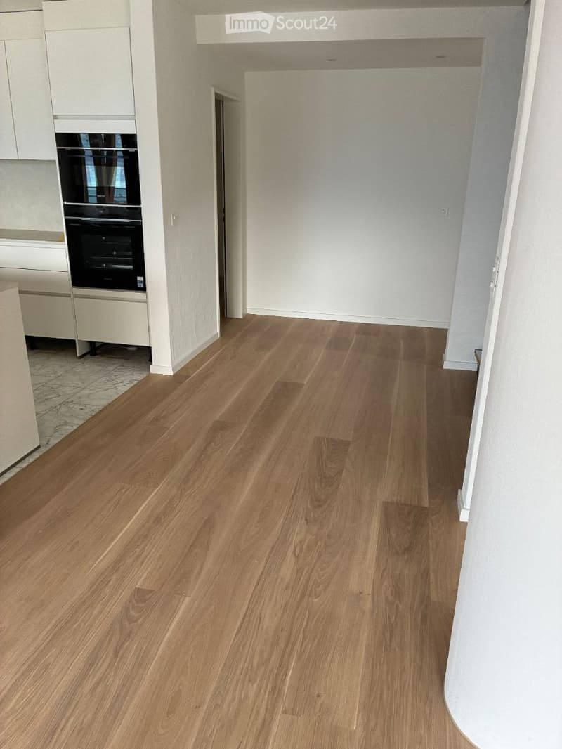 Empty hallway with light wooden flooring and white walls, leading to a kitchen area with white cabinets and a built-in oven.