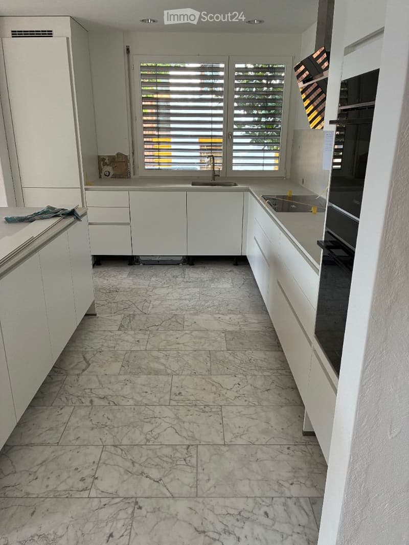 Empty kitchen with white cabinets, marble flooring, and a window with blinds. No appliances or furniture are present.