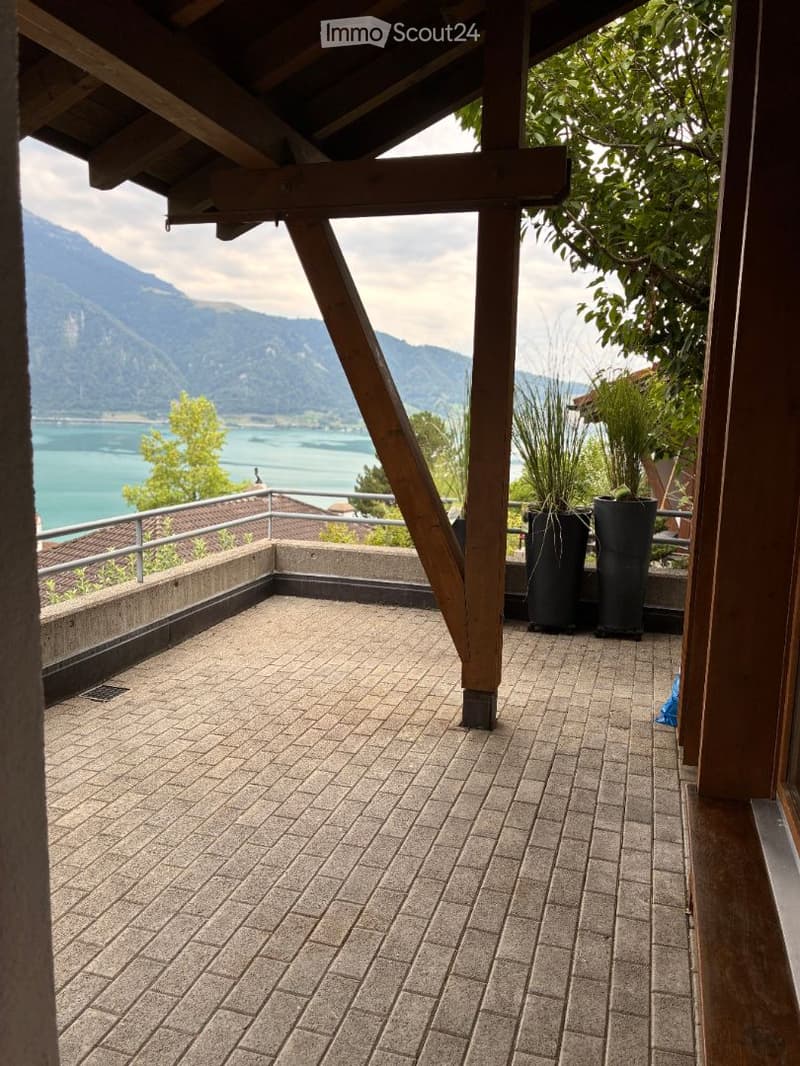 Brick-paved balcony with wooden beams, overlooking a lake and mountains, with potted plants on the edge.