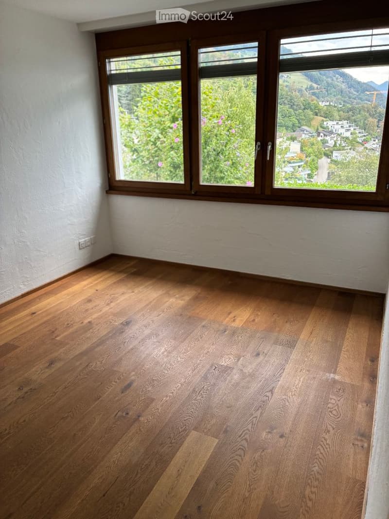 Empty room with wooden flooring and large windows offering a scenic view of greenery and mountains.