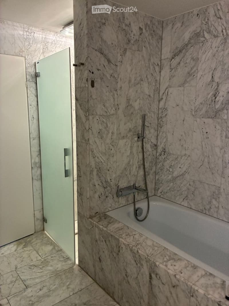 Modern bathroom with marble walls and floor, featuring a bathtub with a handheld shower hose and a glass shower door.