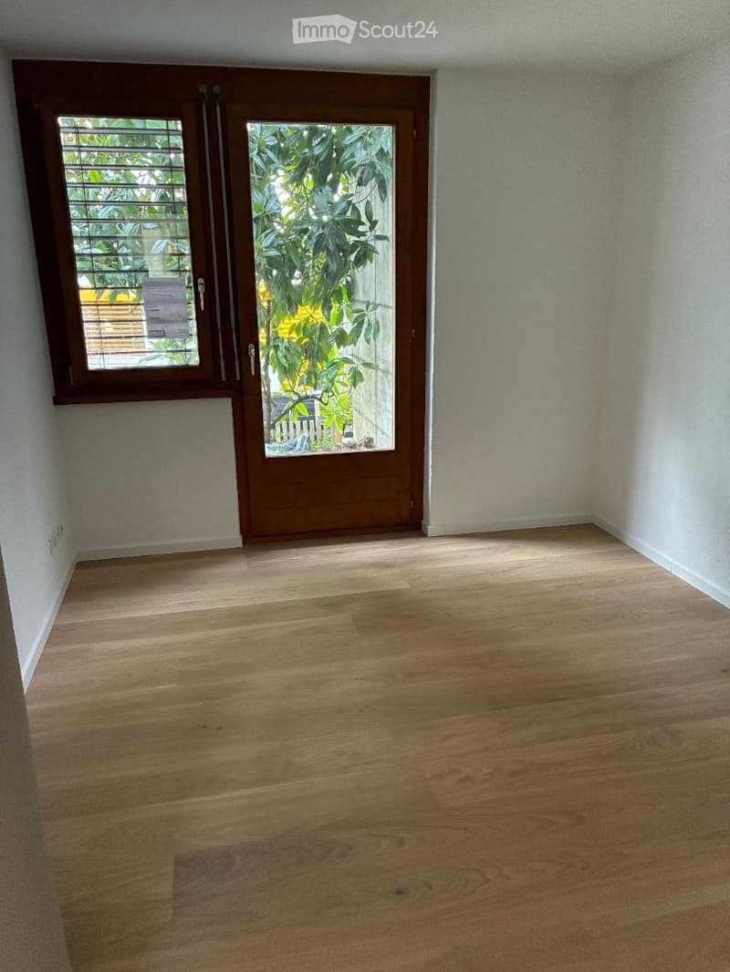 Empty room with wooden flooring, white walls, and a large wooden door leading to a balcony with greenery visible outside.