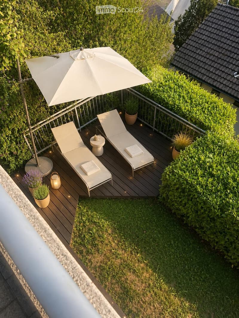 A wooden deck terrace with two lounge chairs, a small table, an umbrella, and potted plants, surrounded by greenery and a metal railing.
