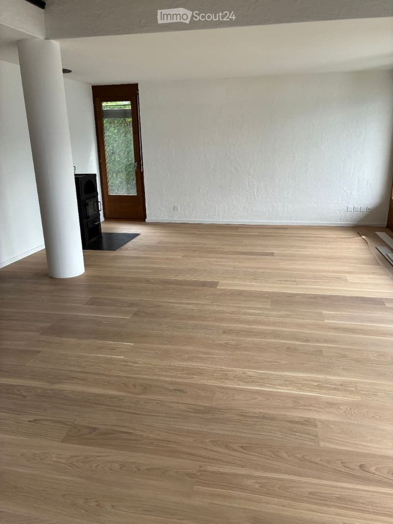 Completely empty room with light wooden flooring, white walls, and a large central column. A wooden door leads to an outdoor area, and a small black fireplace is visible in the corner.