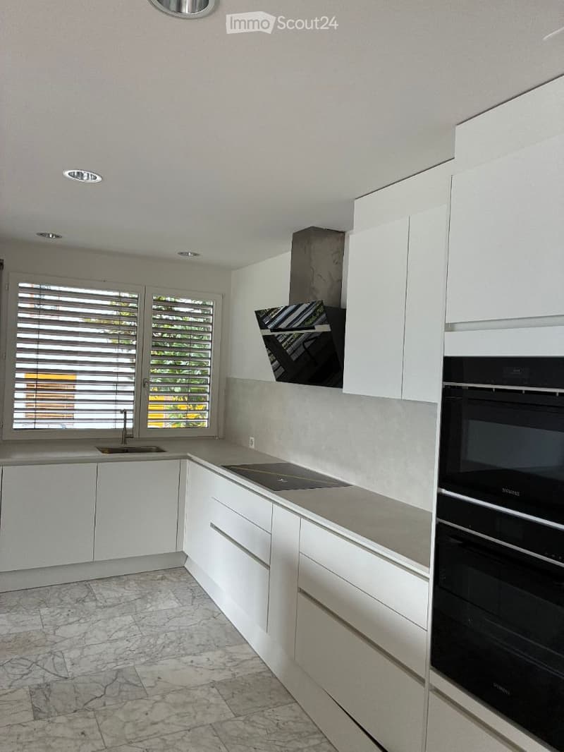 Modern kitchen with white cabinets, marble flooring, and stainless steel appliances including an oven and range hood. Features a large window with blinds and recessed lighting.