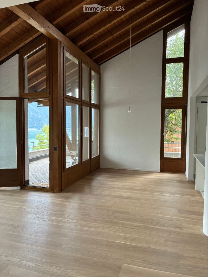 Bright, empty room with wooden flooring, large windows, and exposed wooden beams on the ceiling. The room has a view of a scenic landscape through the windows.