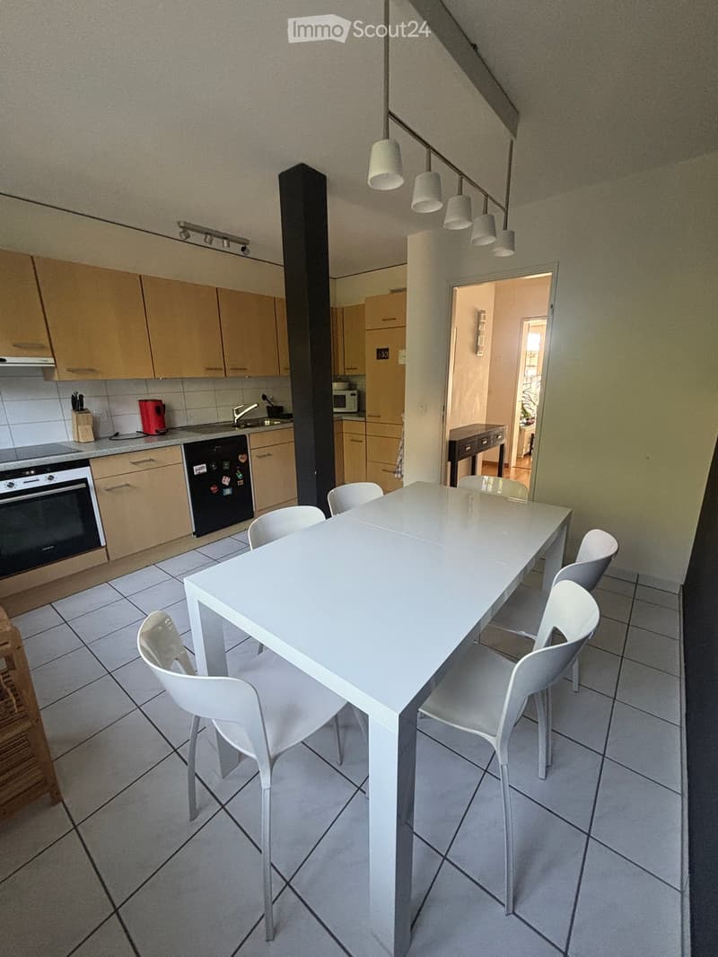 Modern kitchen with light wooden cabinets, white tiled floor, and a long white dining table with six white chairs. Appliances include an oven, refrigerator, and microwave. A small sink with a faucet is visible.