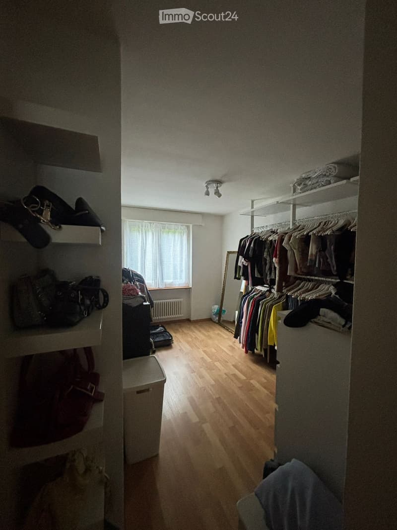 A well-lit, organized walk-in closet with wooden flooring, a large wardrobe filled with hanging clothes, a mirror, and various storage items including bags and a suitcase.