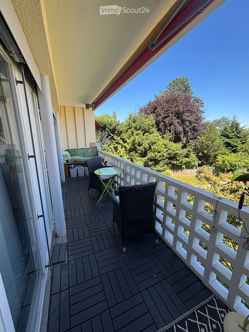 Balcony with a view of trees, featuring a small round table, two wicker chairs, and a green sofa with folded blankets on it.