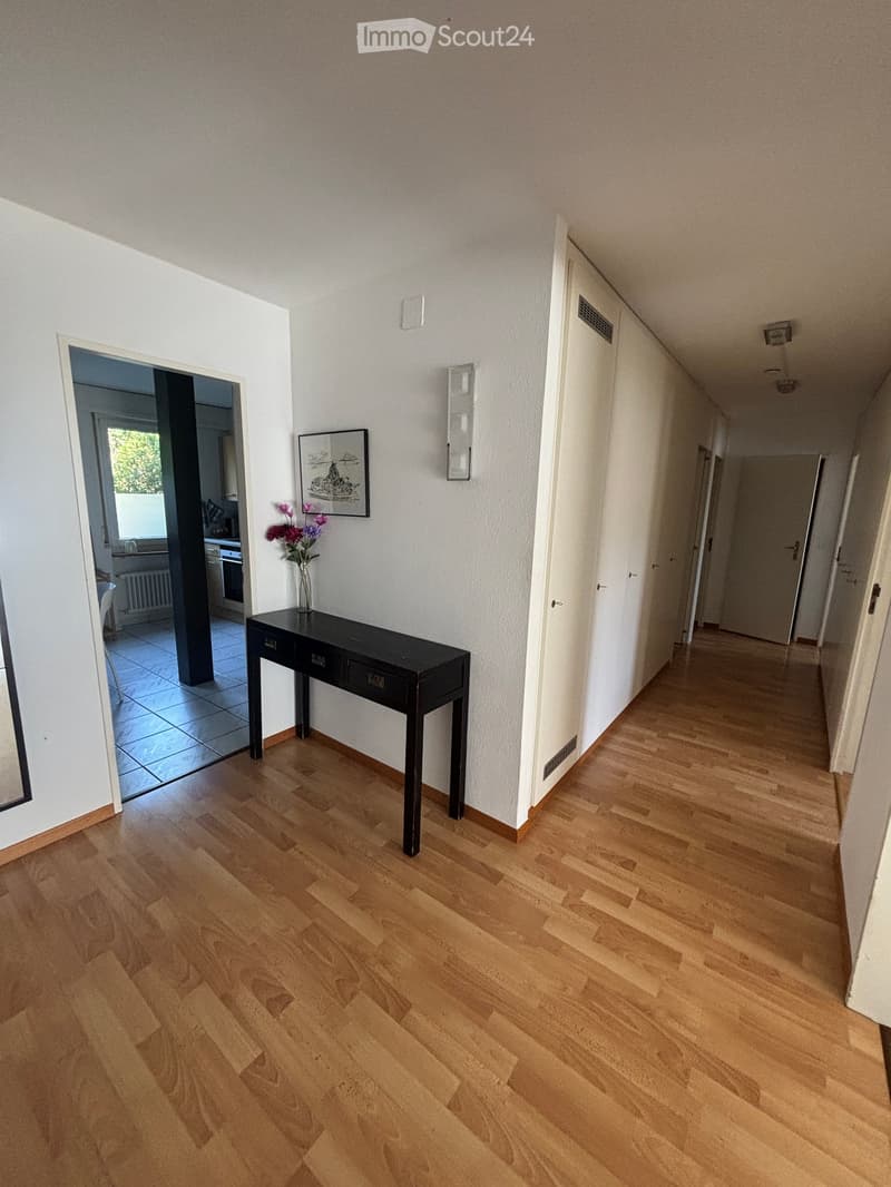 A long, well-lit hallway with wooden flooring, white walls, and several doors leading to different rooms. A small black console table with a vase of flowers is placed against one wall, and a framed picture hangs above it. The hallway has a modern and clea