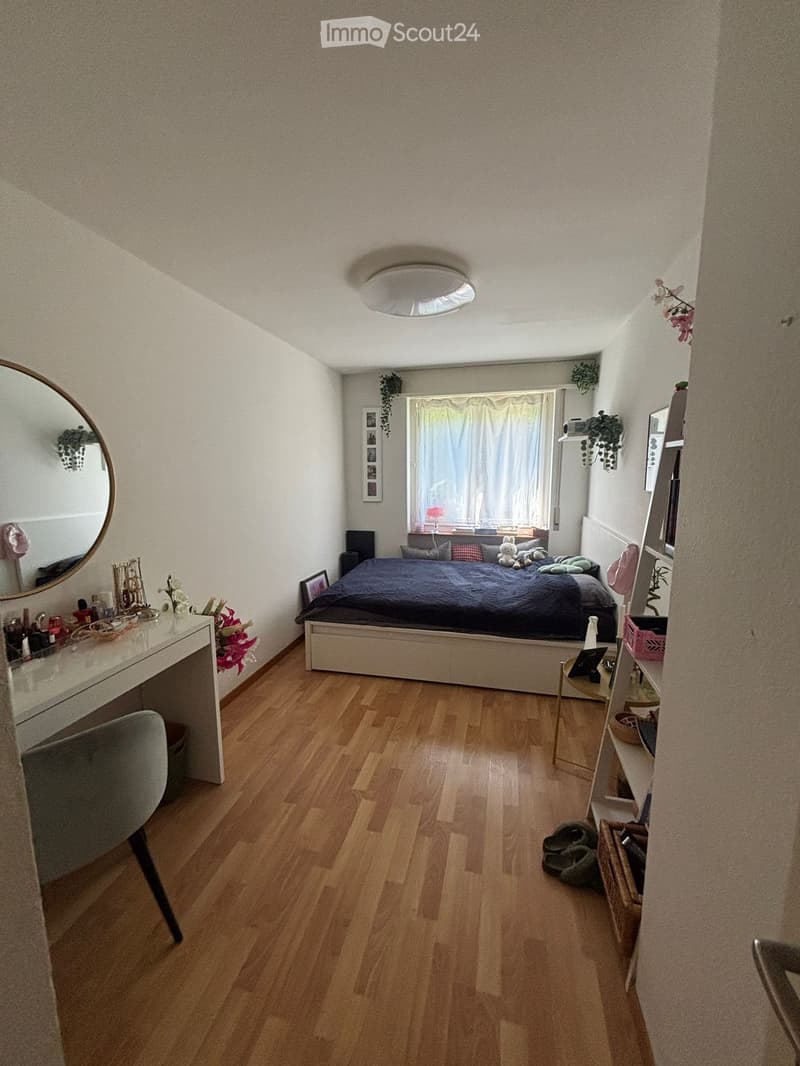 Cozy bedroom with wooden flooring, a bed with dark bedding, a vanity with a mirror, a chair, and a small table with decorative items.