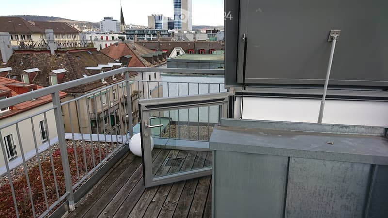 Balcony with wooden decking, metal railing, glass door leading to a small seating area, and a view of the cityscape with rooftops and buildings.