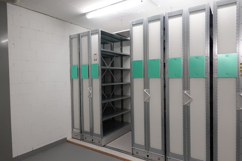 An empty room with white brick walls and gray flooring. There are large metal storage units with green panels and silver handles. The room is well-lit with ceiling lights.