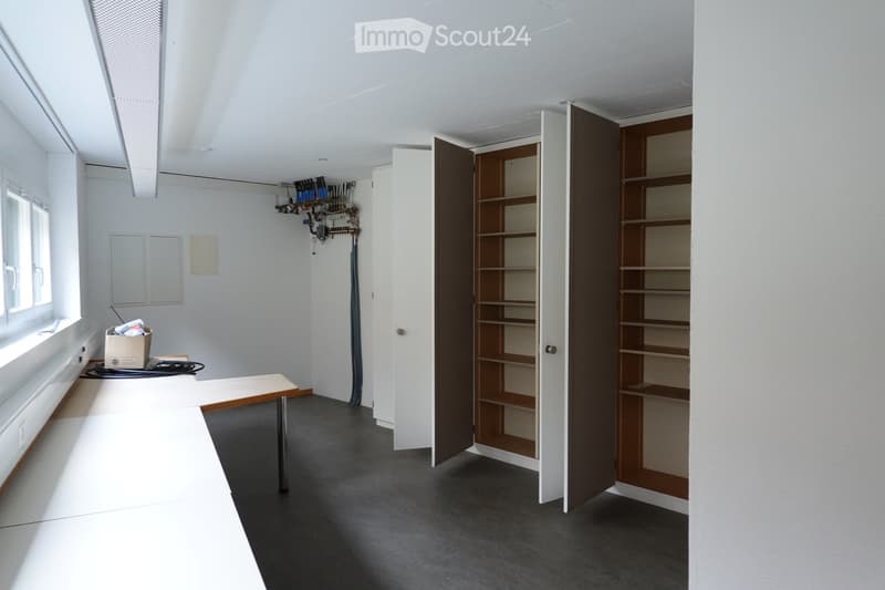An empty room with white walls, gray flooring, and built-in wooden shelves. There is a desk with a box on it and a window with a view outside.