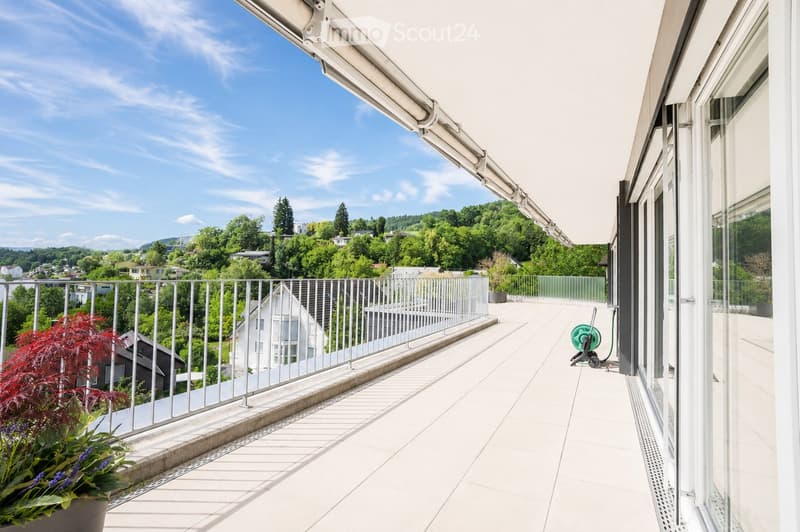 Spacious balcony with white tiled flooring, metal railing, and a retractable awning. Features potted plants and a garden hose reel, overlooking a green, hilly landscape with houses and trees.