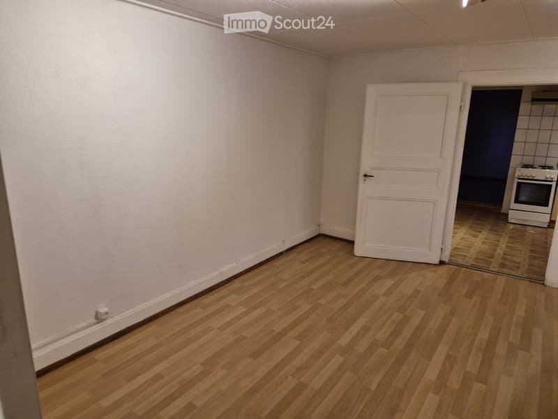 Empty room with light wooden flooring and white walls, featuring a white door leading to another room with a visible oven.