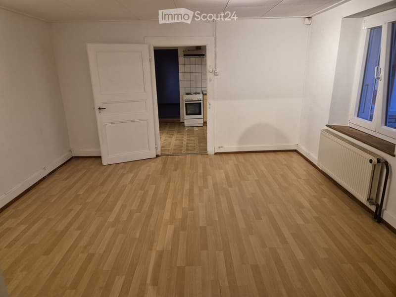 Empty room with wooden flooring, white walls, a white door leading to a kitchen area, and a window with blue glass panes.