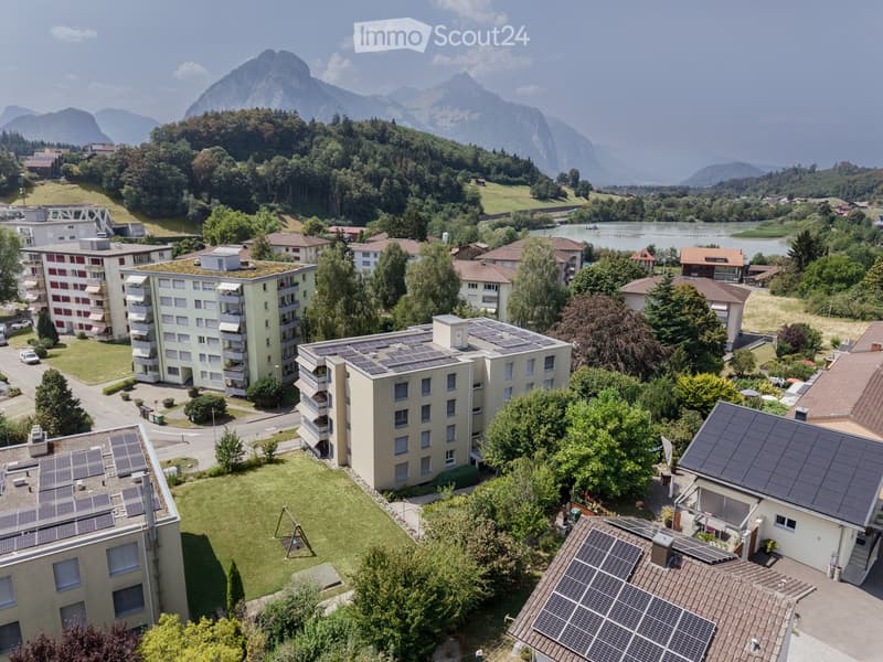 Aerial view of a residential area with multiple buildings, green spaces, and solar panels on rooftops, surrounded by trees and mountains.