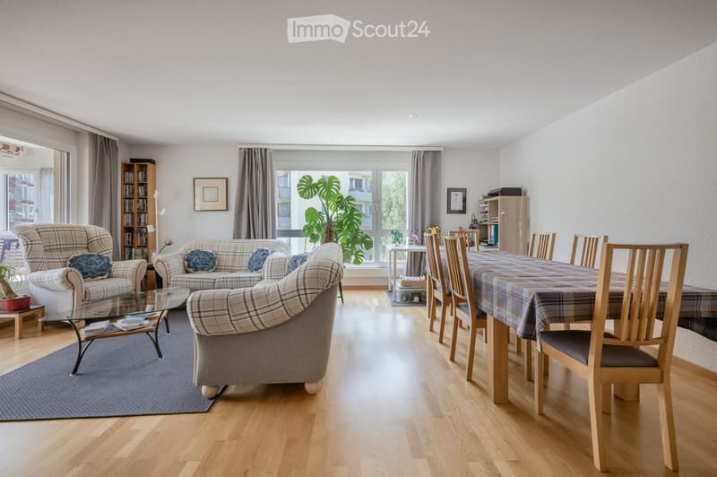 Bright living and dining area with wooden flooring, large windows, plaid sofas, a glass coffee table, a dining table with chairs, and a bookshelf.