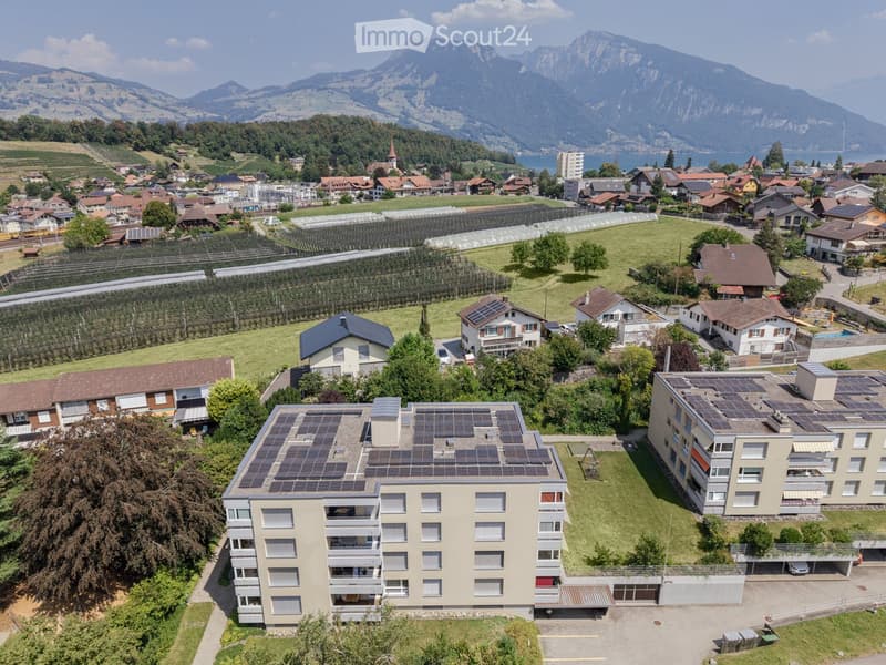 Aerial view of a residential area with multiple buildings, solar panels on rooftops, and surrounding greenery and mountains.