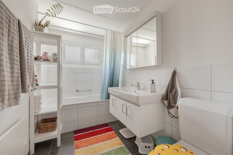 Modern bathroom with white tiles, a sink, mirror, bathtub, toilet, and a shelf with various items. A colorful rug is on the floor, and a towel hangs on the wall.