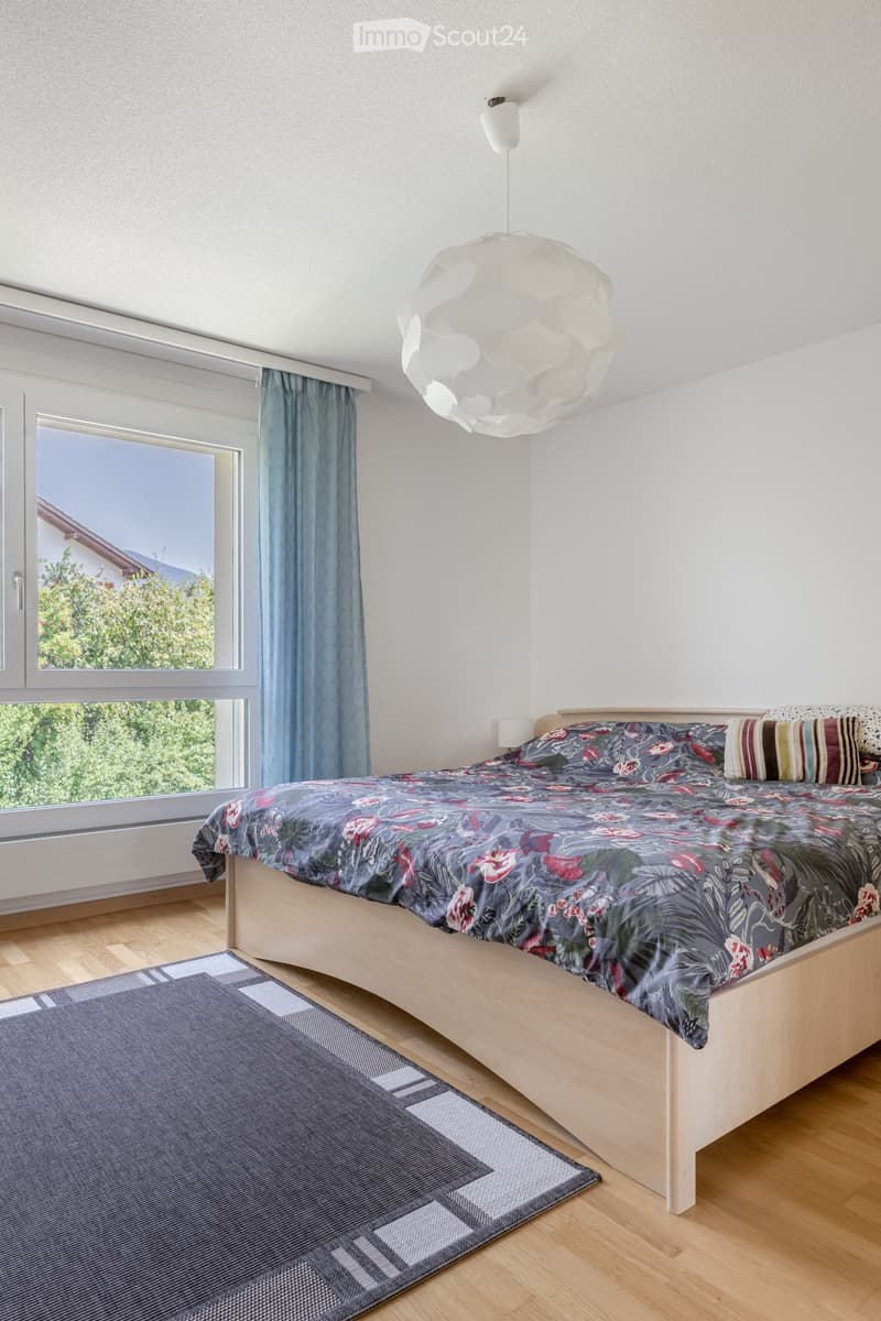 Bright bedroom with wooden flooring, large window with blue curtains, bed with floral duvet and pillows, grey rug, and a modern hanging lamp.