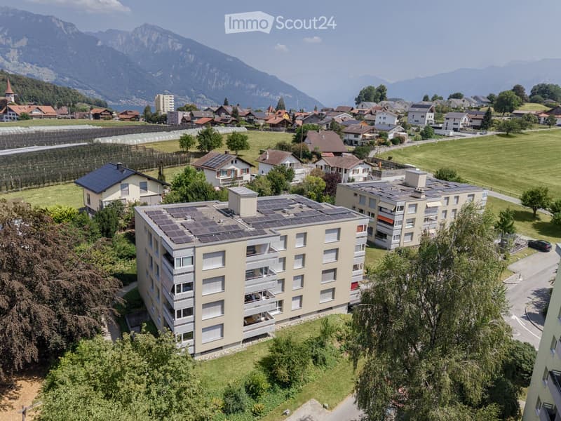 Aerial view of a multi-story residential building with solar panels on the roof, surrounded by greenery and other houses in a scenic valley with mountains in the background.