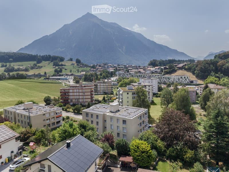 Aerial view of a residential area with multiple apartment buildings, surrounded by green fields and a mountainous backdrop.